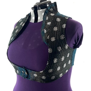May include: A black and silver floral patterned bolero vest with teal trim and a button closure. The vest is displayed on a mannequin with a purple shirt. The vest has a high collar.