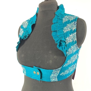 May include: A teal-colored vest with a ruffled collar and decorative floral patterns. The vest has a button closure and is displayed on a black mannequin. The fabric appears to be textured.