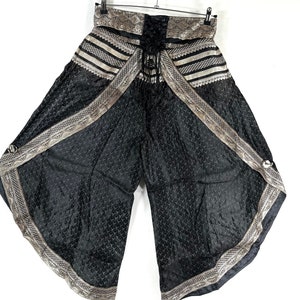 May include: Black palazzo pants with a decorative silver and gold trim. The pants have a lace-up front and a wide, flowing leg design. The fabric has a subtle pattern, and the pants are hanging on a hanger.