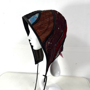May include: A black leather hooded hat with a patchwork design featuring brown, red, and purple fabrics. The hat has a drawstring closure and is decorated with intricate embroidery.