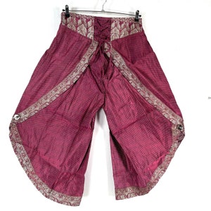May include: Wide-leg, burgundy pants with a patterned design and decorative trim. The pants have a lace-up back and are hanging on a hanger. The fabric has a subtle polka dot pattern. The trim is a metallic silver color.
