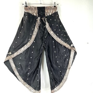 May include: Black palazzo pants with a decorative patterned waistband and trim. The pants feature a lace-up front and a flowing, wide-leg design. Small white floral accents are scattered across the fabric. The trim has a beige and gold pattern.