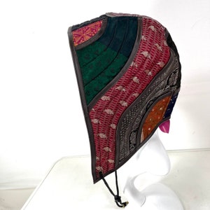 May include: A brown hooded hat with a patchwork design featuring various colours and patterns, including green, red, and gold. The hat has a drawstring closure and is made of a variety of fabrics.