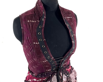 May include: A burgundy vest with a high collar and a lace-up front. The vest features a quilted pattern and black trim with metal eyelets. The bottom of the vest has a layered, patterned fabric with ruffles.