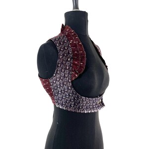 May include: A vintage-style vest with a high collar and a unique pattern. The vest features a dark purple paisley design with silver accents and a ruffled burgundy trim. It is displayed on a black mannequin.