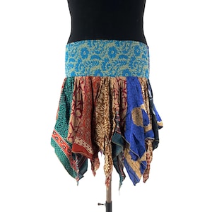 May include: A patchwork skirt with a blue elastic waistband and a variety of patterned fabrics in shades of red, brown, blue, and green. The skirt has an asymmetrical hemline and is displayed on a mannequin.