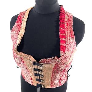 Può includere: Un gilet rosso e oro fantasia con design a collo halter. Il gilet presenta un pannello frontale dorato con cinturini neri e bottoni dorati. I bordi sono rifiniti con un tessuto dorato a balze.