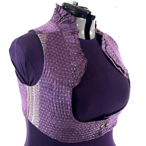May include: A purple bolero jacket with a textured, pleated design and silver accents. The jacket features a high, ruffled collar and fastens with two small buttons. The garment is displayed on a purple mannequin.