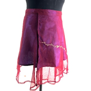 Könnte beinhalten: Ein Wickelrock in Lila- und Magentatönen mit einem transparenten, gerüschten Saum. Der Rock hat ein gesticktes Golddesign und ist aus einem glänzenden, fließenden Stoff gefertigt. Der Rock wird auf einer Schaufensterpuppe präsentiert.