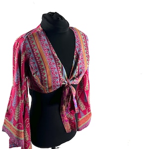 May include: A magenta-coloured cropped top with flared sleeves. It has a V-neck and a tie fastening at the waist. The fabric displays a repeating pattern of orange, blue, and green vertical stripes and floral details.
