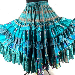 Puede incluir: Falda larga escalonada en tonos turquesa, azul y verde. La falda presenta una cintura con cordón y múltiples capas de tela con volantes. La tela tiene un diseño estampado con elementos florales y abstractos, creando una estética bohemia.