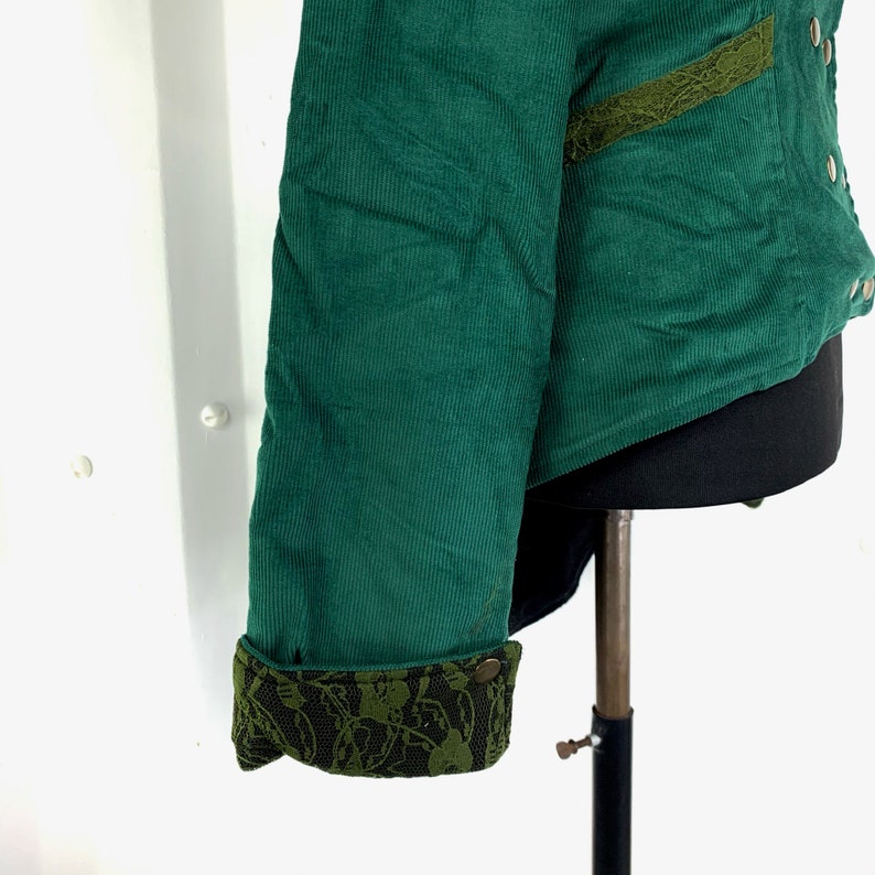May include: A green corduroy jacket with a lace trim on the sleeves and a pocket. The jacket has a black lining and a button closure.