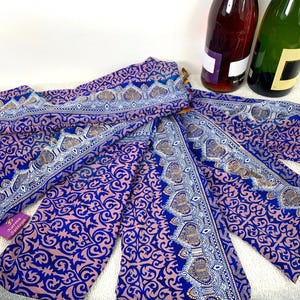 Peut inclure: Écharpe à motifs bleus et roses avec un motif de bordure détaillé. L'écharpe est drapée, mettant en valeur son motif complexe. Deux bouteilles de vin sont en arrière-plan. L'écharpe porte le texte "Karma Sutra" sur une étiquette.