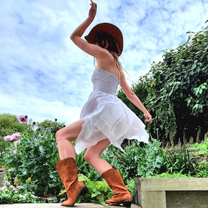 May include: A woman wearing a white dress and a brown cowboy hat is twirling in a field. She is wearing brown cowboy boots.