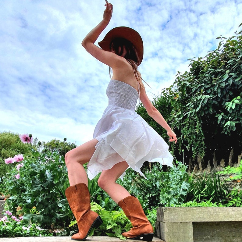 May include: A woman wearing a white dress and a brown cowboy hat is twirling in a field. She is wearing brown cowboy boots.