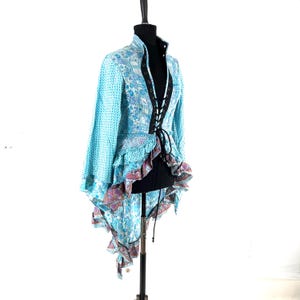 May include: A light blue, long-sleeved jacket with a high collar and a lace-up front. The jacket features a mix of patterned fabrics, including a floral design and a ruffled hem with a paisley print. The jacket is displayed on a black mannequin.