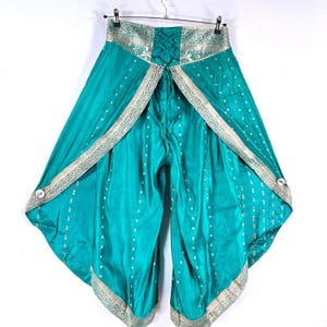 May include: Teal-colored palazzo pants with a decorative silver trim and small white polka dots. The pants have a unique design with an open front and a lace-up back. The fabric appears to be a silky material, and the pants are hanging on a hanger.