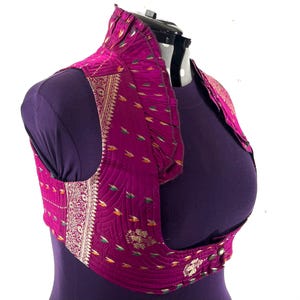 May include: A magenta-colored vest with a high collar and decorative gold and silver trim. The vest features a quilted pattern with small, colorful leaf-like designs. It has a button closure and is displayed on a mannequin.