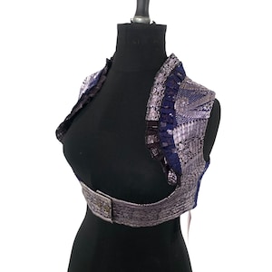 May include: A cropped bolero jacket in shades of blue and silver, displayed on a black mannequin. The jacket features intricate patterns, a ruffled collar, and a buckle closure at the waist. The fabric has a metallic sheen.