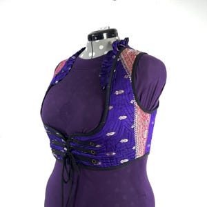 May include: A purple vest with a black trim and a lace-up front. The vest has a patterned design with silver accents and a pink and red border. The vest is displayed on a mannequin.