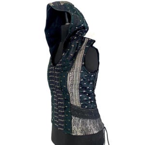 May include: A sleeveless hooded vest in dark green and black featuring intricate silver paisley patterns, vertical striped panels, and black lace trim. The garment includes a side lace-up closure with black cord ties and a structured hood.