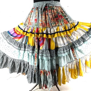 May include: A multi-layered, bohemian-style skirt with a drawstring waist. The skirt features a patchwork design with various patterns, including floral, striped, and abstract prints. Colors include yellow, blue, red, and black. The skirt has multiple ruffles and a flowing silhouette.