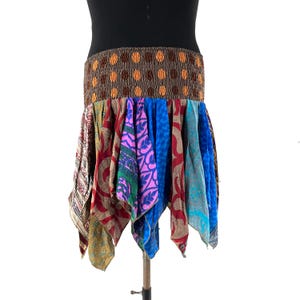 May include: A colorful, patchwork skirt with a brown elastic waistband featuring orange polka dots. The skirt is made of various patterned fabrics in shades of red, blue, green, and purple. The skirt is displayed on a mannequin.