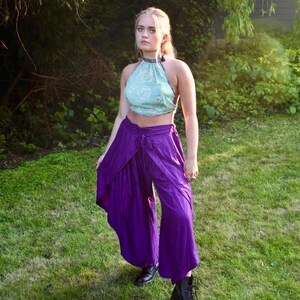 May include: A halter top with a light blue floral pattern and purple palazzo pants. The pants have a wrap-style design. The person is wearing black boots. The outfit is set against a backdrop of green grass and foliage.