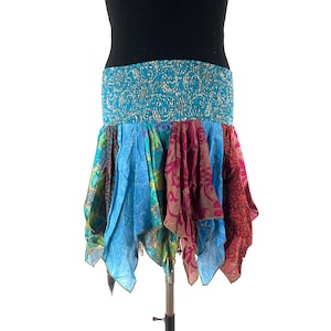 Può includere: Una gonna colorata in stile bohémien con una fascia elastica turchese. La gonna presenta pannelli di tessuto vibrante nelle tonalità di blu, verde e rosso, creando un orlo asimmetrico a strati. La gonna è esposta su un manichino.