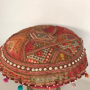 May include: A round, multi-colored, embroidered floor cushion with a shell trim and tassels. The cushion is sitting on a woven base.
