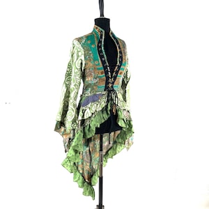 Op de afbeelding: Een groen en goudkleurig jasje met een hoge kraag en lange mouwen. De jas heeft een veter aan de voorkant, ruches en een asymmetrische zoom. De stof heeft een patchwork-ontwerp met tinten groen, goud en bruin.