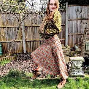 May include: A woman wearing a green and brown patterned jacket and a long, orange and brown patterned skirt. She is standing in a garden with a wooden fence in the background.