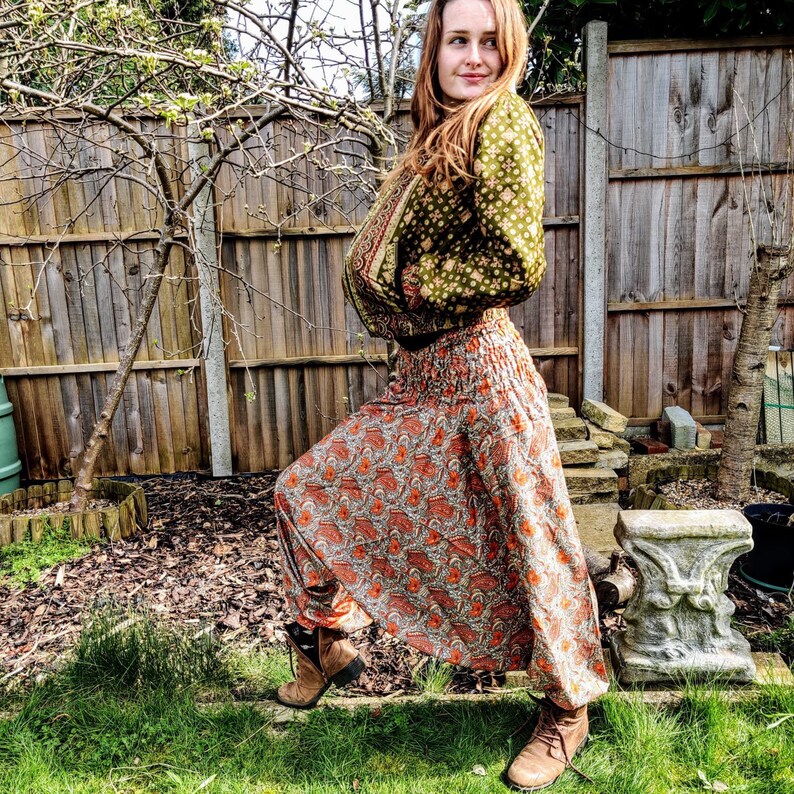 May include: A woman wearing a green and brown patterned jacket and a long, orange and brown patterned skirt. She is standing in a garden with a wooden fence in the background.