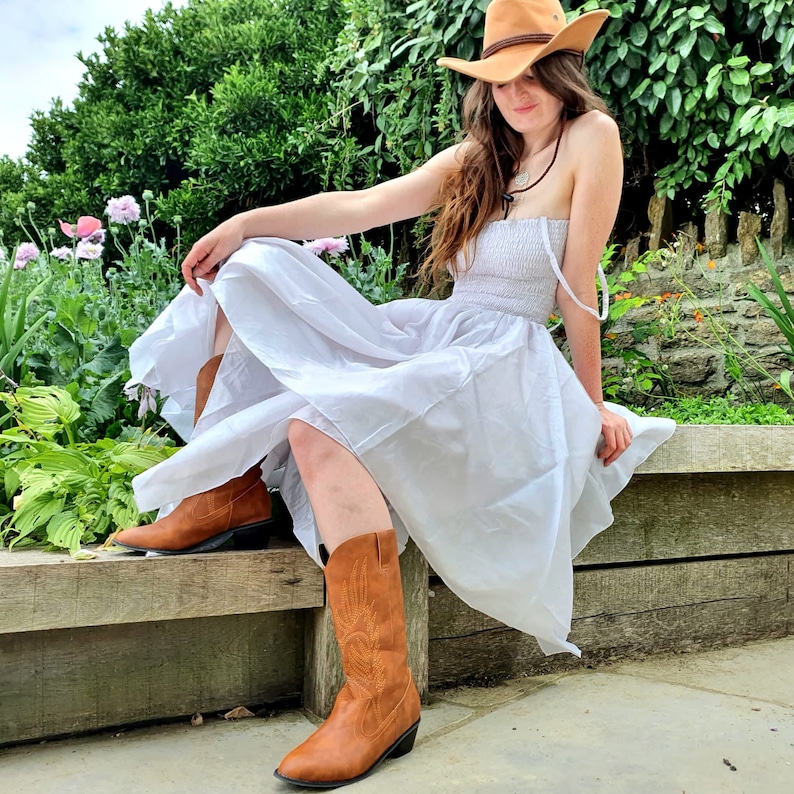 May include: A woman wearing a white sundress and brown cowboy boots, with a brown cowboy hat on her head. She is sitting on a wooden bench with green plants in the background.