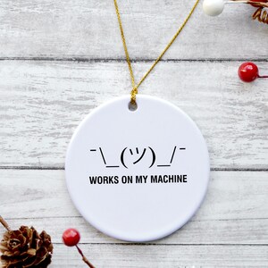 May include: A round white ceramic ornament featuring a black printed shrug emoticon and the text "WORKS ON MY MACHINE" in a sans-serif font. The ornament is suspended by a thin gold string, creating a simple and clear design for display.