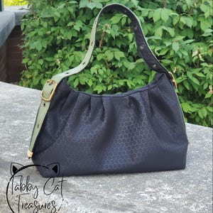 May include: Black handbag with a honeycomb pattern. The bag has a light green strap with gold-colored hardware. The bag is sitting on a concrete surface with green foliage in the background. The text "Tabby Cat Treasures" is at the bottom.