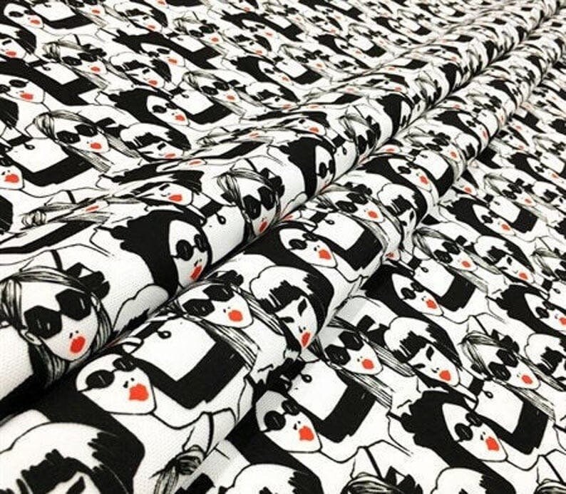 May include: White fabric with a repeating pattern of stylized faces in black and white. Each face has sunglasses and red lips. The fabric is folded, showing the pattern's repeating design.