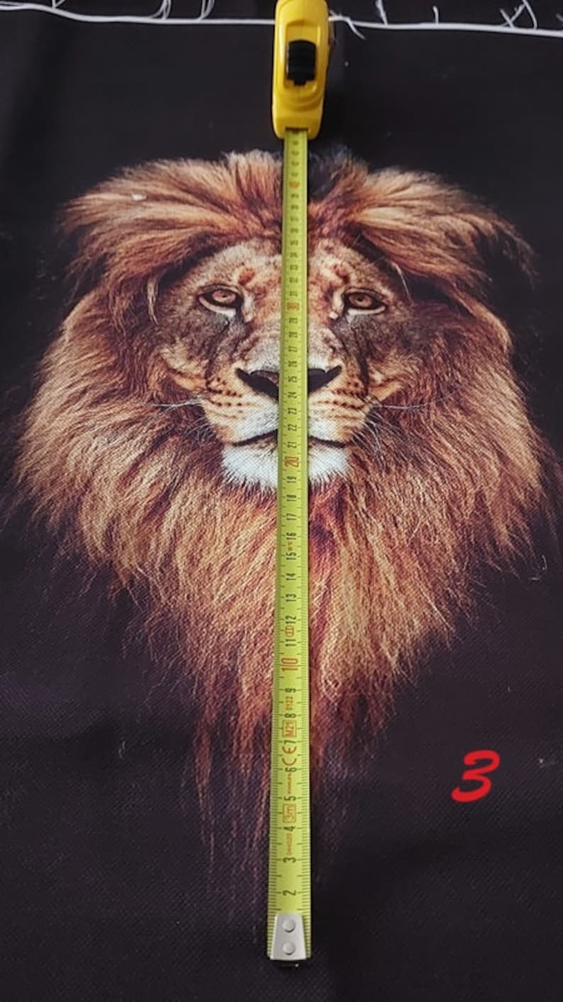 May include: A black fabric panel featuring a detailed, realistic lion head print in shades of brown, gold, and tan. A yellow measuring tape is placed vertically over the lion's face, showing a measurement of approximately 32 centimetres in length.