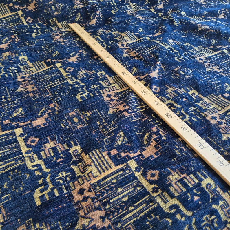 May include: A deep blue fabric featuring a complex, repeating geometric pattern in shades of light tan and gold. A wooden ruler is placed diagonally across the textile, showing measurements in centimetres up to 75 cm for scale and reference.