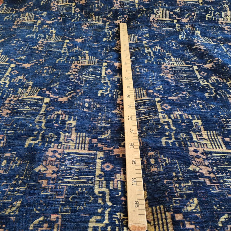 May include: A deep blue fabric featuring a repeating geometric pattern in light tan and cream. A wooden ruler is placed vertically on the textile, showing measurements in centimetres ranging from 20 cm to 95 cm along its length.