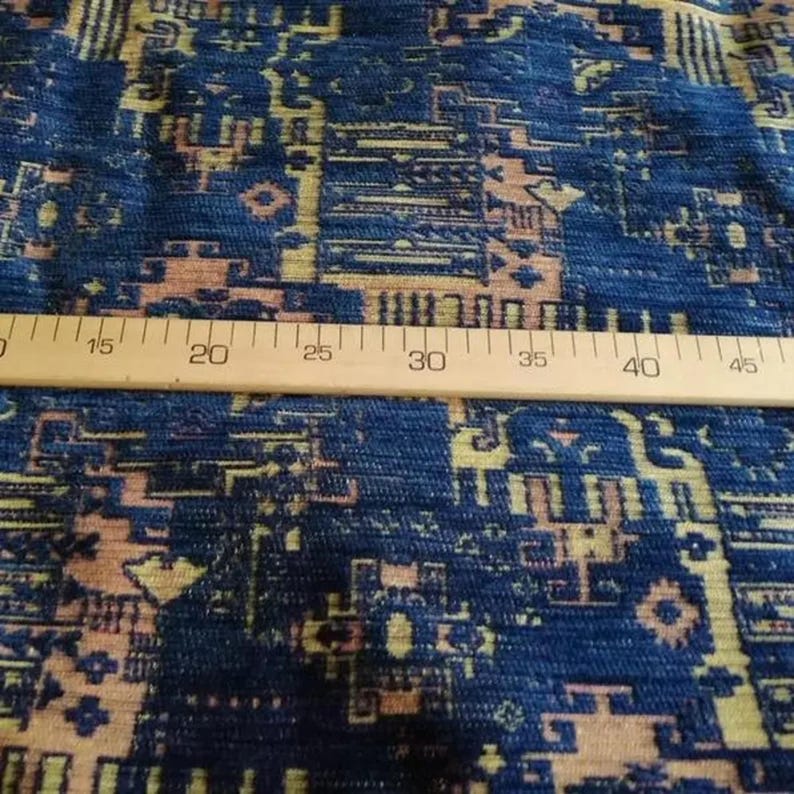 May include: A blue fabric featuring a geometric pattern in shades of tan and cream. A wooden ruler is placed horizontally across the textile, showing measurements from 15 to 45 cm, indicating the scale of the intricate woven design.