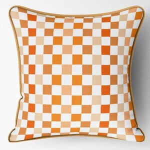 K&ouml;nnte beinhalten: Quadratisches Zierkissen mit Schachbrettmuster in Orange-, Wei&szlig;- und Beiget&ouml;nen. Das Kissen hat einen beigen Rand. Wohnaccessoire.