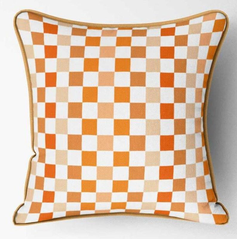 K&ouml;nnte beinhalten: Quadratisches Zierkissen mit Schachbrettmuster in Orange-, Wei&szlig;- und Beiget&ouml;nen. Das Kissen hat einen beigen Rand. Wohnaccessoire.