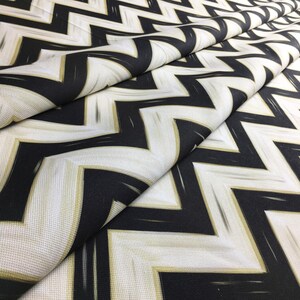 May include: Close-up of a folded fabric with a bold zigzag pattern. The design features black and white chevrons with subtle gold accents. The fabric has a textured appearance, suggesting a durable material suitable for various applications.