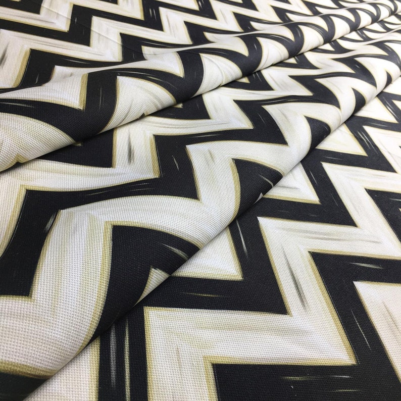 May include: Close-up of a folded fabric with a bold zigzag pattern. The design features black and white chevrons with subtle gold accents. The fabric has a textured appearance, suggesting a durable material suitable for various applications.