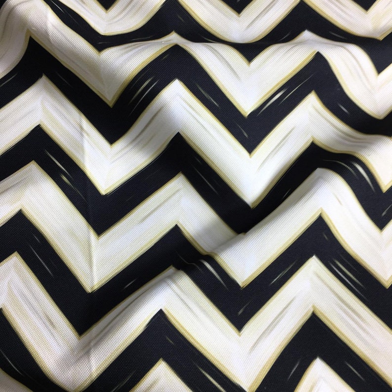 May include: Close-up of a fabric with a bold chevron pattern. The design features alternating black and white zigzags, with gold brushstroke accents. The fabric has a slightly textured appearance, with soft folds.