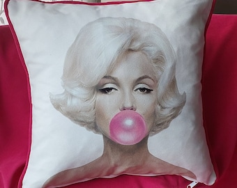 May include: Square decorative pillow featuring a portrait of a woman with blonde hair blowing a pink bubble. The pillow has a white background with a pink border and a zipper.