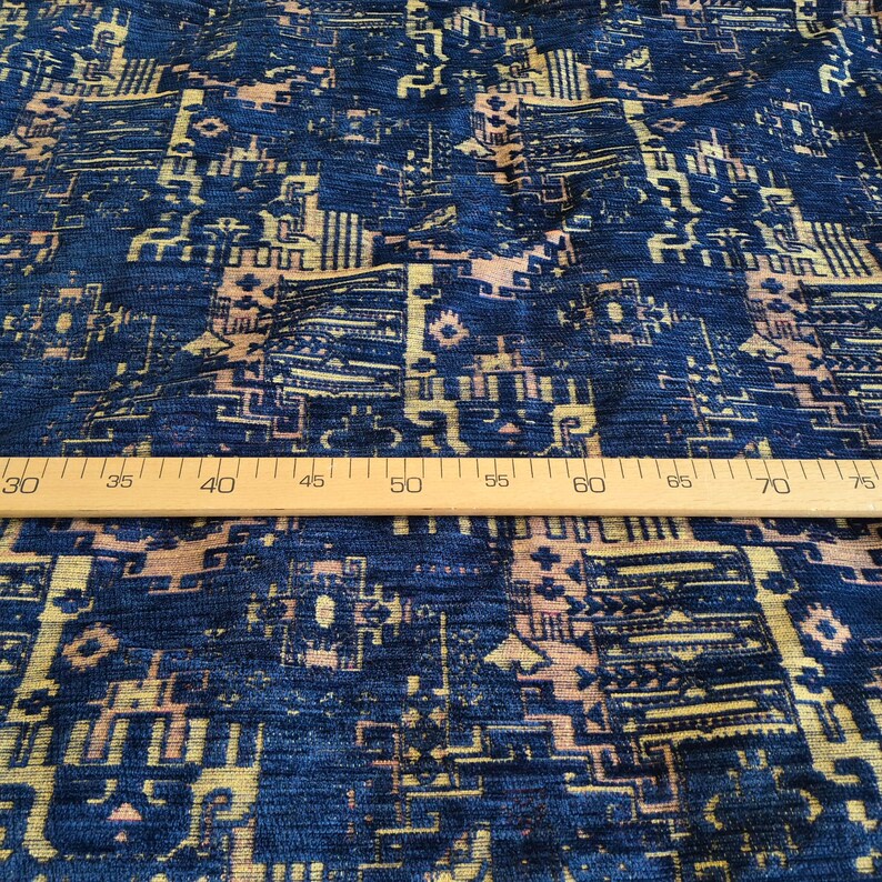 May include: A dark blue fabric featuring a repeating geometric pattern in shades of gold and light peach. A wooden ruler is placed horizontally across the centre of the textile, showing measurements from 30 to 75 centimetres for scale.