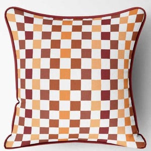 K&ouml;nnte beinhalten: Quadratisches Zierkissen mit einem karierten Muster in Wei&szlig;, Orange, Braun und Burgunderrot. Das Kissen hat einen burgunderroten Rand. Dieses Kissen eignet sich f&uuml;r die Inneneinrichtung.