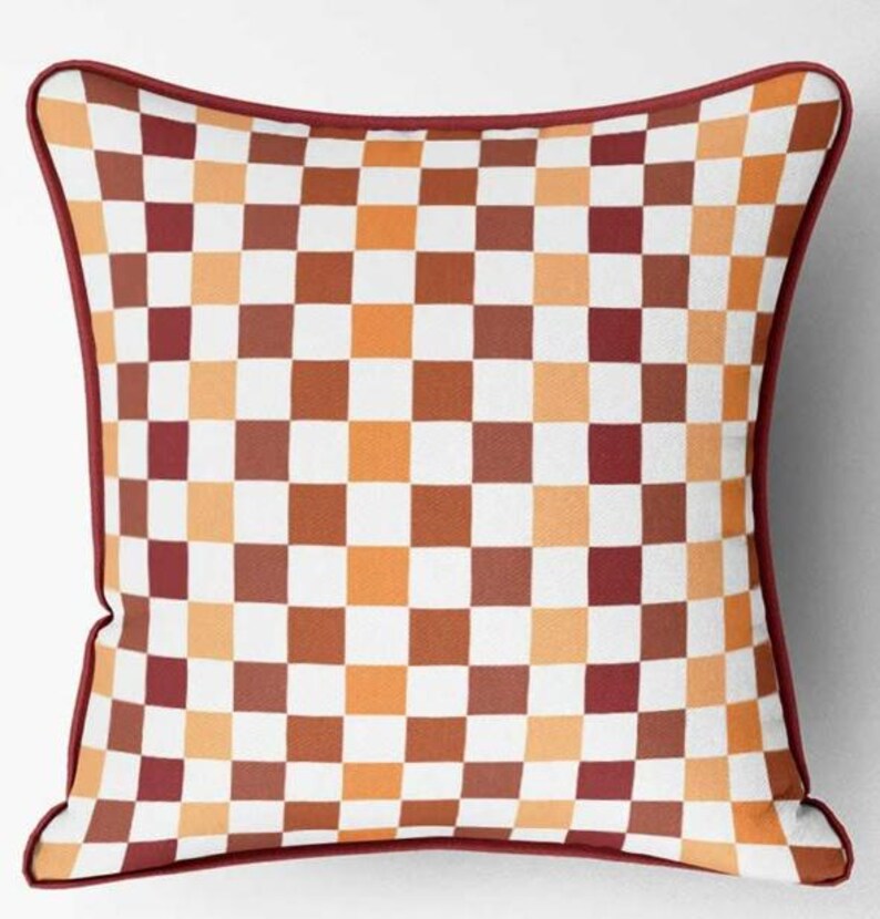 K&ouml;nnte beinhalten: Quadratisches Zierkissen mit einem karierten Muster in Wei&szlig;, Orange, Braun und Burgunderrot. Das Kissen hat einen burgunderroten Rand. Dieses Kissen eignet sich f&uuml;r die Inneneinrichtung.